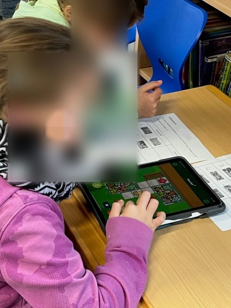 Hier sind Zwei Mädchen zu sehen die zusammen an einem vor ihnen liegendem Tablet arbeitet, auf dem ein Level der besagten Bee-Bot App gepiet wird.