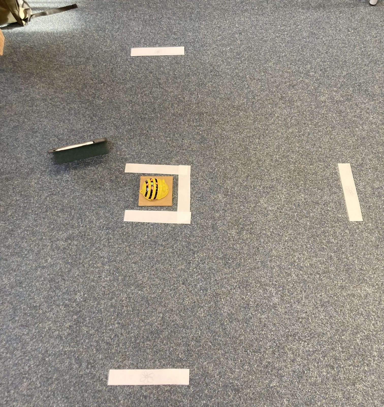 Station 2
Es ist erneut ein Bee-Bot zu sehen, welcher in der Mitte des Bildes auf dem Boden steht. Orthogonal dazu sind in drei Richtungen jeweils ein Papierschnipse mit einer Blüte drauf ebenfalls auf den Boden gelegt. Neben dem Roboter steht ein Tablet.
