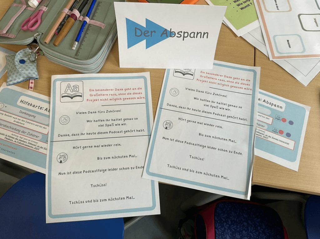 Das Bild zeigt einen Arbeitsplatz in einer Schule. Auf dem Tisch ist eine Station mit verschiedenem Material, wie beispielsweise Formulierungshilfen, für das Erstellen eines Abspanns für einen Podcast aufgebaut.