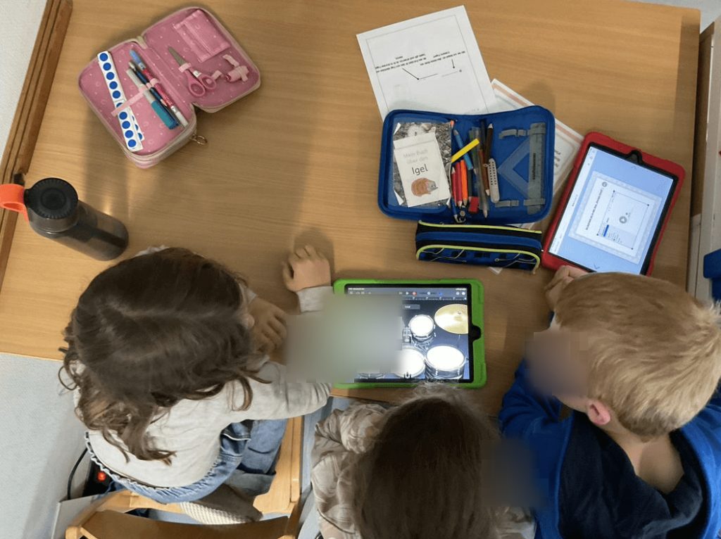 Das Bild zeigt drei Kinder aus der Vogelperspektive, die gemeinsam an einem Tisch arbeiten und auf ein IPad schauen, auf dem ein Schlagzeug abgebildet ist.