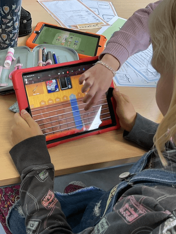 Das Foto zeigt eine Nahaufname von zwei Mädchen von hinten, die zusammen ein Ipad mit der App GarageBand bedienen. Eine Schülerin hält das IPad fest, die andere spielt auf digitalen Gitarrenseiten.