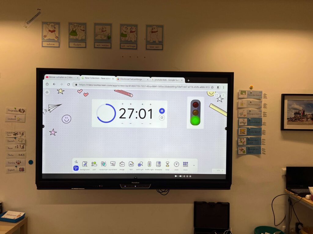 Dieses Foto zeigt ein großes, an der Wand montiertes Smartboard in einem Grundschulklassenzimmer. Auf dem Bildschirm ist die Anwendung „ClassroomScreen“ geöffnet, die einen Timer bei etwa 27 Minuten, eine Ampel, die auf Grün steht, und verschiedene Funktions-Icons am unteren Rand zeigt. Über dem Bildschirm hängen bebilderte Klassenregeln mit Eisbären-Motiven (z. B. „zuhören“, „leise sein“), und links sowie rechts vom Board sind laminierte Kärtchen für den Tagesablauf und die Klassendienste an der Wand befestigt.