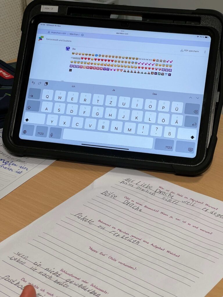 Die Aufnahme zeigt einen hölzernen Schülerschreibtisch, auf dem ein Tablet in einer robusten schwarzen Schutzhülle und ein Arbeitsblatt liegen. Auf dem Tablet ist eine Lernplattform (fobizz) zu sehen, in deren Textfeld eine lange Reihe verschiedener Emojis eingegeben wurde. Im Vordergrund liegt ein handschriftlich ausgefülltes Arbeitsblatt auf Deutsch zum Thema Märchen, auf dem Fragen zu Helden, Bösewichten (hier: „böser Drache“) und Aufgaben innerhalb der Geschichte beantwortet wurden.