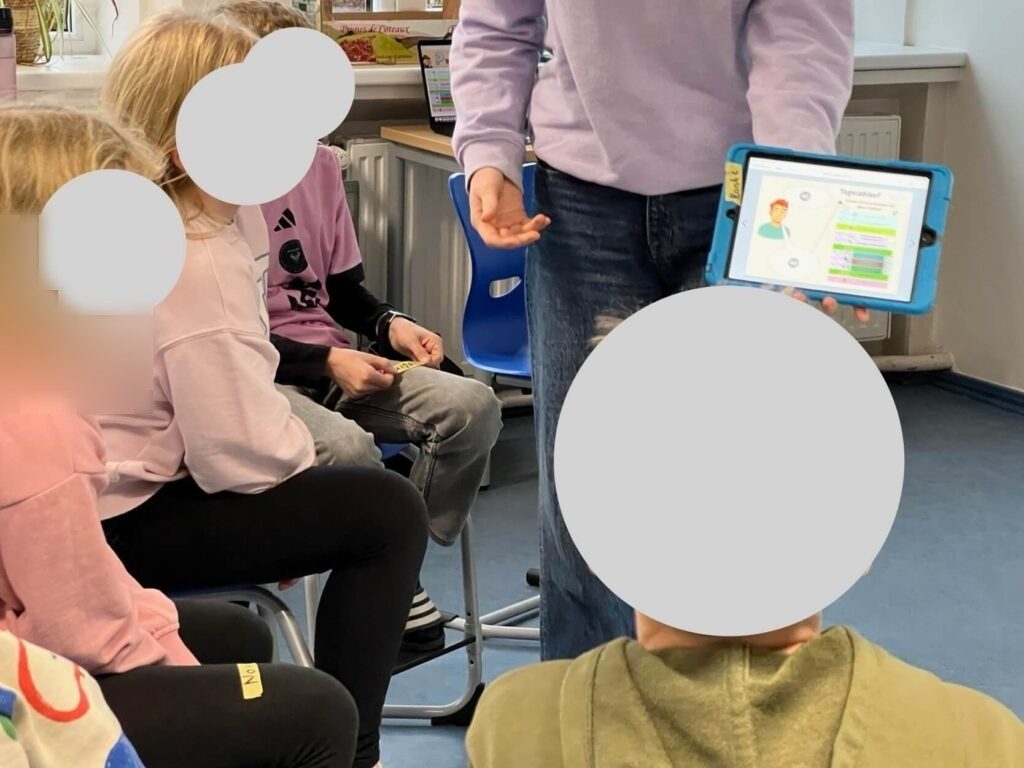 Die Schüler:innen sitzen im Sitzkreis während die Studentin mit dem digitalen Bookcreator herumgeht und die entsprechenden Funktionen erklärt.