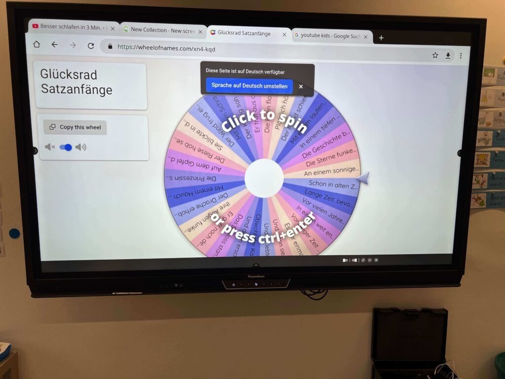Auf dem digitalen Whiteboard im Klassenzimmer ist die Webseite „wheelofnames.com“ mit einem bunten, interaktiven Glücksrad zum Thema „Satzanfänge“ geöffnet. Die einzelnen Segmente des Rades enthalten verschiedene Textbausteine wie „Vor vielen Jahren...“, „Plötzlich hörte er...“ oder „An einem sonnigen...“. In der Mitte des Rades erscheint die Aufforderung „Click to spin“, während im Browser verschiedene Tabs zu pädagogischen Themen und YouTube Kids sichtbar sind.