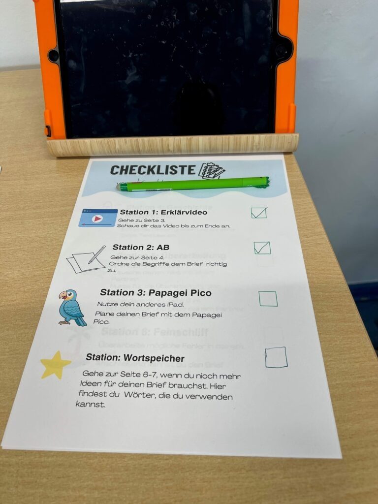 Das Foto zeigt ein Lernarrangement auf einem hellen Holztisch, das der Selbstorganisation während eines Projekttages dient. Im oberen Bildteil steht ein Tablet mit einer auffälligen orangefarbenen Schutzhülle in einer Halterung aus Bambus; der Bildschirm ist ausgeschaltet. Direkt davor liegt eine gedruckte „Checkliste“ im A4-Format, auf der ein grüner Stift abgelegt wurde. Die Liste führt verschiedene Lernstationen auf, die jeweils mit einem Symbol und kurzen Anweisungen versehen sind: Station 1 (Erklärvideo): Mit einem Play-Symbol markiert und bereits mit einem grünen Häkchen als erledigt gekennzeichnet. Station 2 (AB): Mit einem Blatt-und-Stift-Symbol markiert, ebenfalls bereits abgehakt. Station 3 (Papagei Pico): Illustriert mit dem blauen Papageien Pico; hier geht es um die Planung eines Briefes. Das Kontrollkästchen ist noch leer. Station: Wortspeicher: Mit einem gelben Stern markiert, dient als Hilfestellung für Ideen und Formulierungen. Das Bild illustriert die methodische Strukturierung des Unterrichts, bei der digitale Medien (Tablet) und analoge Organisationshilfen (Checkliste) kombiniert werden, um den Lernfortschritt für die Schülerinnen und Schüler sichtbar zu machen.