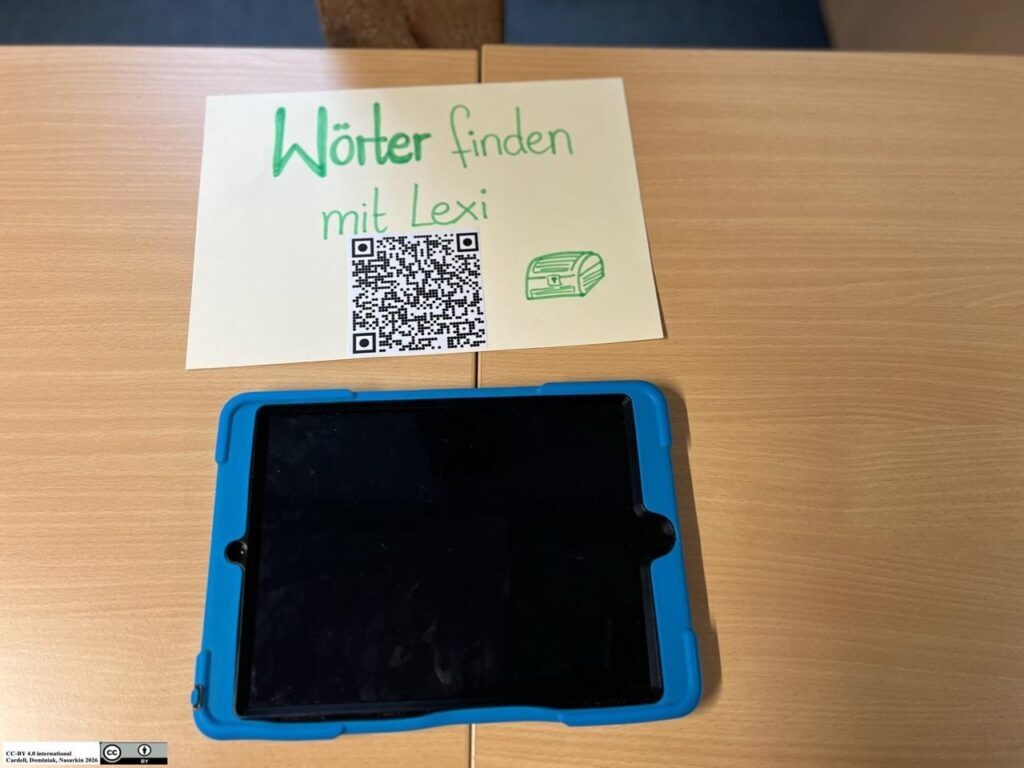 Das Foto zeigt eine Lernstation auf einer hellen Holzoberfläche, bestehend aus einem Hinweisschild und einem Tablet. Im oberen Bereich liegt ein gelbes Blatt Papier, auf dem in grüner Handschrift der Titel „Wörter finden mit Lexi“ steht. Unter dem Text ist ein quadratischer QR-Code abgedruckt, daneben befindet sich eine kleine Illustration einer Schatzkiste. Direkt unterhalb des Blattes liegt ein flach platziertes Tablet in einer robusten, leuchtend blauen Schutzhülle. Der Bildschirm des Geräts ist schwarz und ausgeschaltet. Die gesamte Szenerie wirkt wie eine vorbereitete digitale Lernaufgabe für Kinder in einer Schule oder Kindertagesstätte.