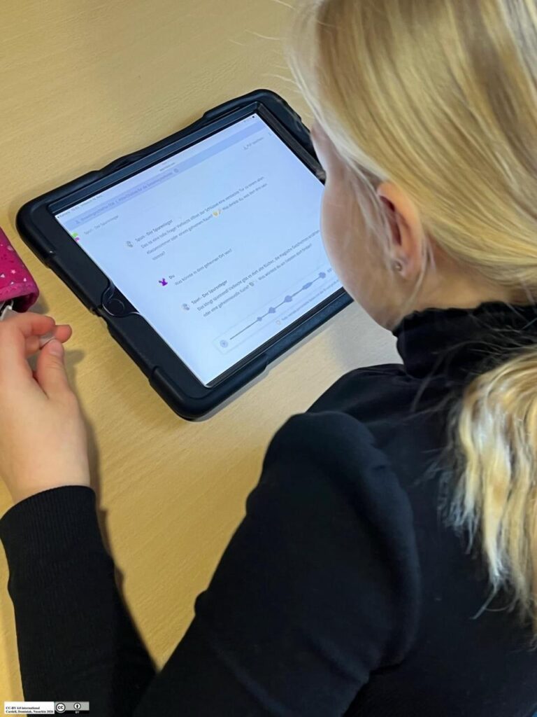 Ein Kind mit blondem Haar sitzt an einem Tisch und schaut auf ein Tablet. Auf dem Bildschirm ist ein Chatbot zu sehen, mit dem das Kind in Interaktion tritt.