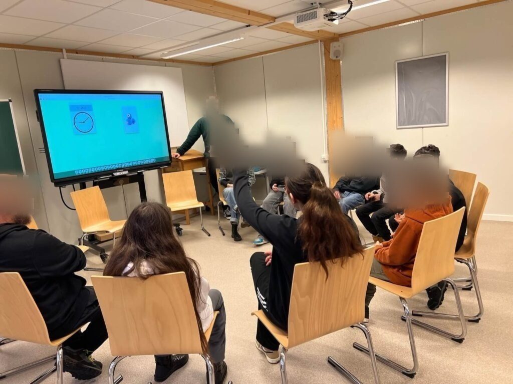 Schüler:innen sitzen im Stuhlkreis vor dem Smartboard. Eine Schülerin meldet sich. Auf dem Smartboards ist eine Uhr und eine Abbildung des Dash-Roboters.