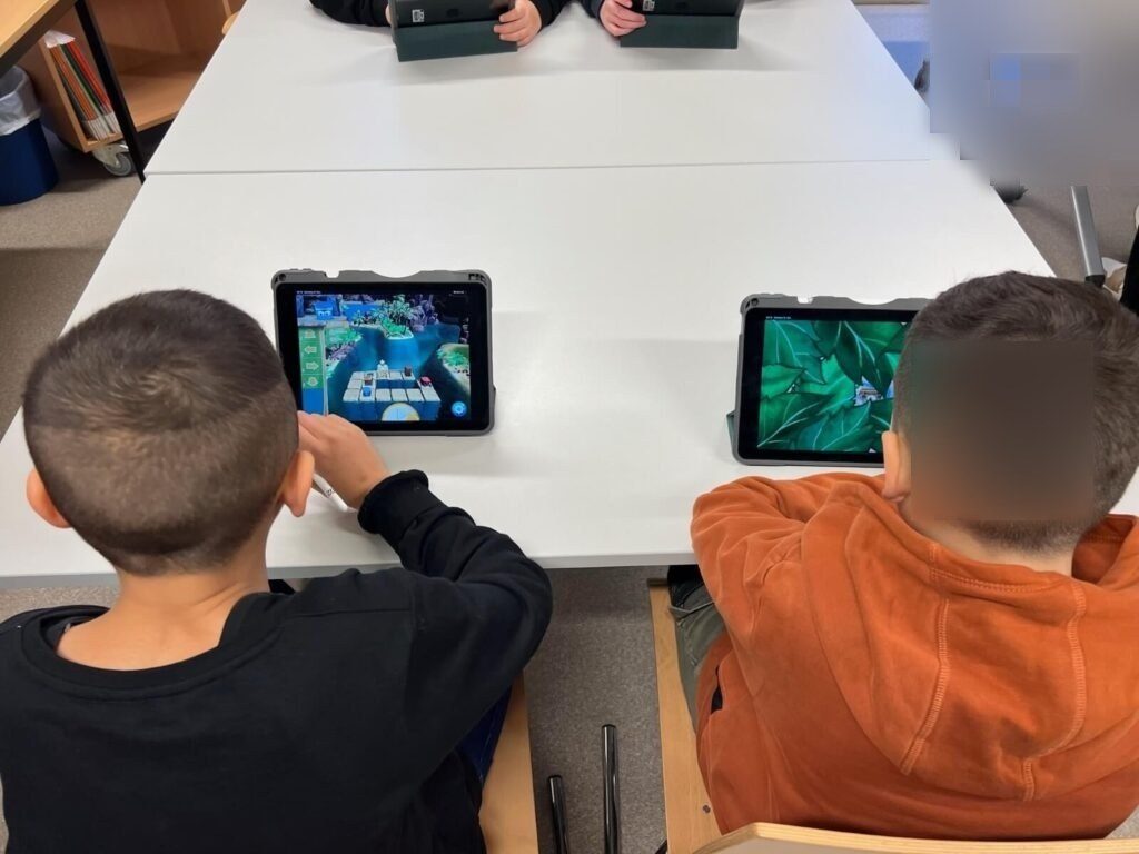 Zwei Schüler sitzen vor jeweils einem iPad. Auf den iPads ist die App Box-Island geöffnet. Sie sitzen an einem Tisch gegenüber von zwei anderen Schüler:innen, die auch jeweils an einem iPad arbeiten.