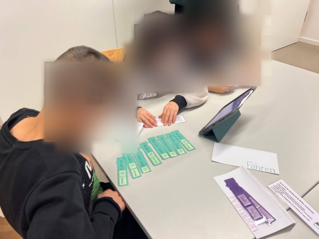 An einem Tisch sitzen drei Kinder. Sie arbeiten mit einem iPad. Vor ihnen auf dem Tisch verteilt liegen die Blockly-Papier-Elemente aneinandergereiht. Daneben liegen geöffnete Umschläge