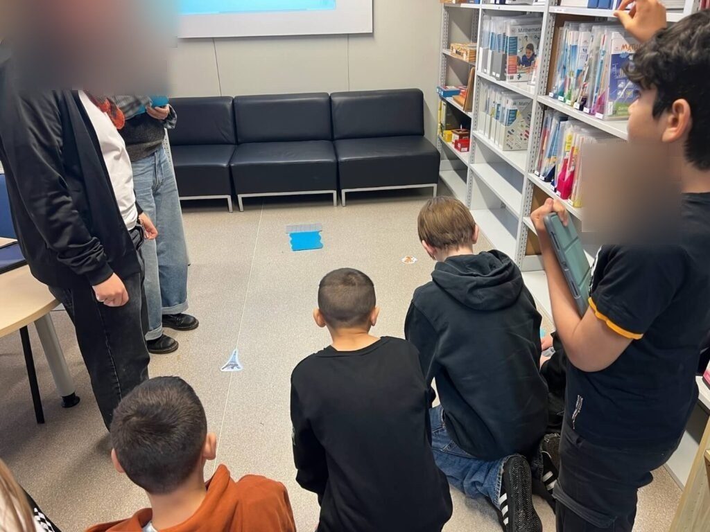 Fünf Kinder sitzen auf dem Boden. Links von ihnen stehen zwei Lehrpersonen. Vor ihnen liegen Papierelemente, wie der Fluss und der Eiffelturm.
