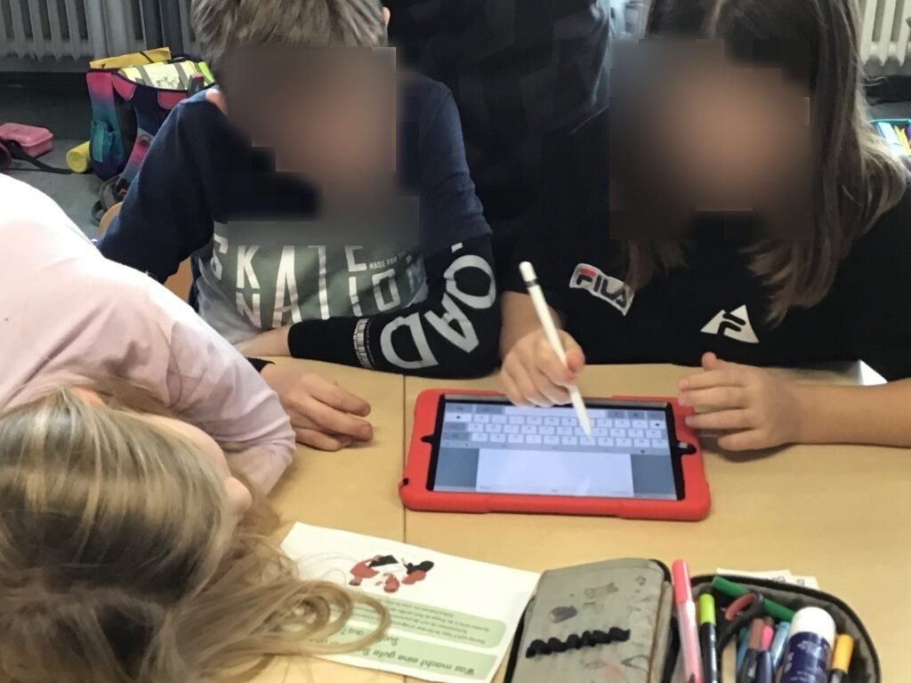 Auf dem Bild zu sehen sind Schüler:innen, die gemeinsam an der Gruppenarbeit arbeiten. Eine von ihnen hat einen Apple Pencil in der Hand und schreibt tippt damit etwas auf der iPad Tastatur. Die anderen Schüler:innen schauen ebenfalls gespannt auf das iPad.