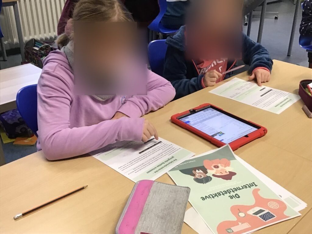 Auf dem Bild sind zwei Schüler:innen zu sehen, die ebenfalls an der Stationsarbeit arbeiten. Zwischen ihnen liegt ein iPad und sie zeigen mit dem Finger auf die Aufgabe, die sie bearbeiten.