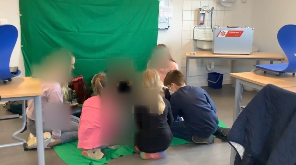 Kinder und Studierende sitzen vor dem Greenscreen und schauen auf das ipad