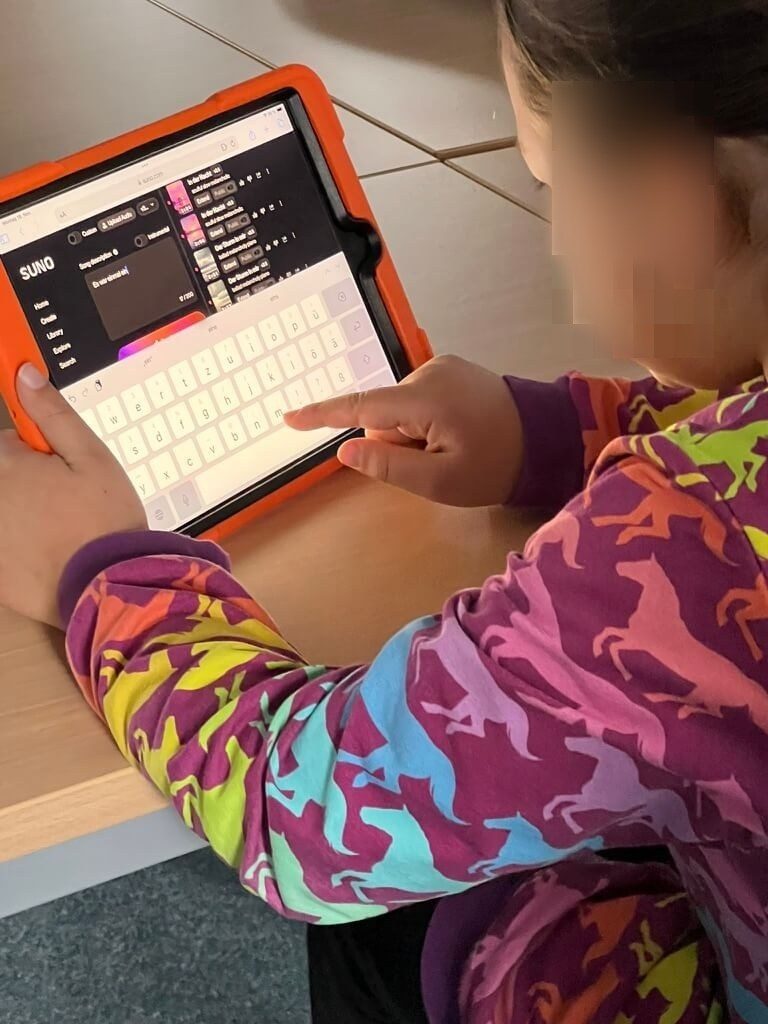 Auf diesem Bild ist ein kleines Mädchen mit einem Bunten Pullover zu sehen, die an einem Schreibtisch sitzt. Auf dem Tisch steht ein IPad, auf dem die Website Suno geöffnet ist. Man kann erkennen, wie das Kind etwas in die Tastatur, welche auf dem IPad eingeblendet ist eintippt.