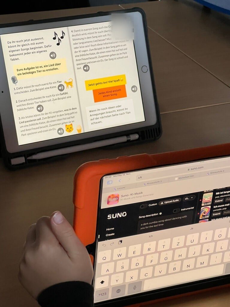 Auf dem Bild sind zwei IPads zu sehen. Auf dem einen ist ein E-Book geöffnet, wo eine Anleitung zu sehen ist, wie man mit Hilfe der Website Suno einen Song erstellt. Das zweite IPad ist näher dran. Dort sieht man die geöffnete Website Suno. Dieses IPad wird von einer Kinderhand festgehalten.