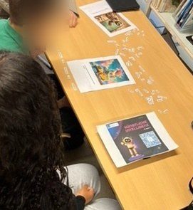 Mehrere Schüler:innen sitzen an einem Holztisch in einer Bibliothek oder einem Klassenraum und arbeiten gemeinsam an einer Aufgabe. Auf dem Tisch liegen ausgedruckte Bilder, darunter eines mit einer KI-generierten Illustration sowie ein weiteres mit dem Titel „Künstliche Intelligenz“. Daneben sind viele kleine ausgeschnittene Wörter oder Begriffe verstreut, die vermutlich für eine Bildbeschreibung oder eine Zuordnungsaufgabe genutzt werden. Die Schüler:innen sind konzentriert, lehnen sich nach vorne und betrachten die Materialien. Im Hintergrund sind Bücherregale sichtbar, was auf eine Lernumgebung hindeutet.