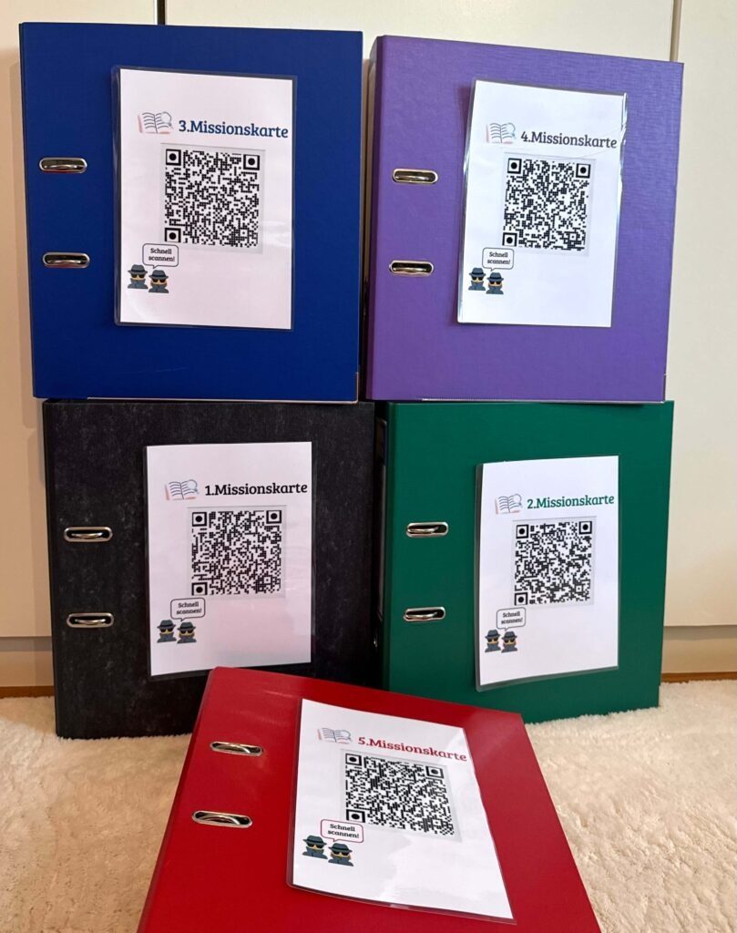 Die fünf Ordner in den Farben rot, blau, schwarz, grün und lila. Jeder Ordner ist mit der entsprechenden Missionskarte (als QR-Code) beklebt.