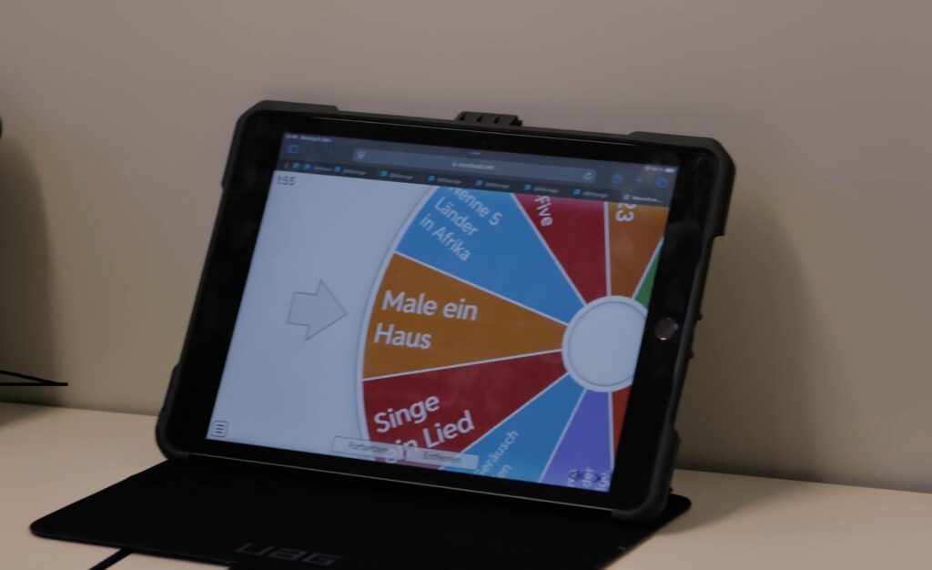 Auf einem iPad ist das digitale Glücksrad zu sehen. Es hat bei der Aufgabe "Male ein Haus" angehalten.
