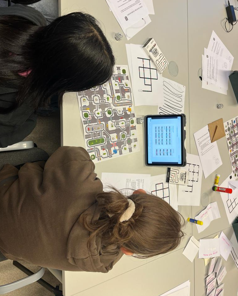Auf diesem Bild sieht man Schüler:innen, die eine Verkehrssituation mit den Ozobots nachstellen. Es liegen verschiedene Materialien auf dem Tisch.