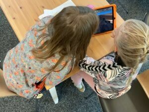 Zwei Mädchen sitzen an einem Tisch, die Kamera fotografiert sie von hinten, ihre Gesichter sind nicht sichtbar. Sie sitzen an einem Tisch und schauen auf ein iPad mit organebner Hülle.
