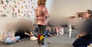 eine Gruppe von Kindern und zwei Erwachsenen sitzen in einem Kreis. Ein Kind steht in der Mitte und hält etwas in der Hand. Die anderen Kindern und die Erwachsenen schauen größtenteils das Kind in der Mitte an, einige schauen in die Gegend.