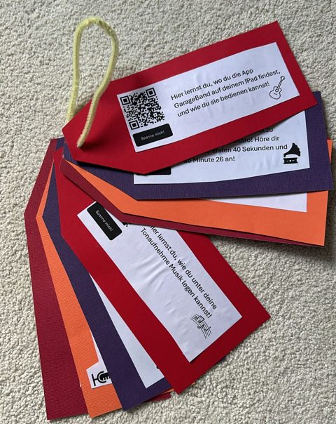 Dieses Bild zeigt mehrere farbige Karten, die an einem gelben Band befestigt sind. Jede Karte hat ein weißes Etikett, auf dem Text und ein QR-Code abgebildet sind. Die Karten sind in verschiedenen Farben, darunter rot, orange und lila. Auf den Etiketten stehen Anweisungen zur Verwendung der App GarageBand auf einem iPad sowie weitere Hilfestellungen zur Audioaufnahme. Neben dem Text befinden sich kleine Symbole, die jeweils zur Erklärung der Aufgaben dienen.