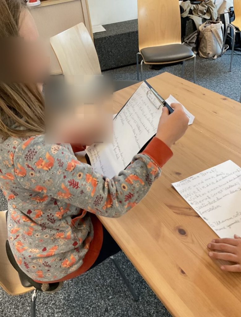 Ein Mädchen sitzt an einem Tisch und liest von einem Blatt Papier, das sie vor sich hält. Auf dem Tisch liegen weitere Blätter, das Mädchen übt die aufgeschriebenen Fragen.