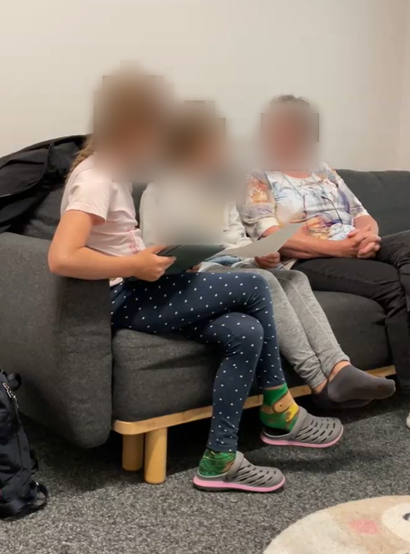 Zwei junge Mädchen sitzen auf einem grauen Sofa neben einer älteren Frau. Die Mädchen halten Tablets und Papierblätter in den Händen und unterhalten sich mit der Frau, die freundlich lächelt. Die Szene zeigt eine entspannte Atmosphäre, in der ein intergenerationales Interview stattfindet.
