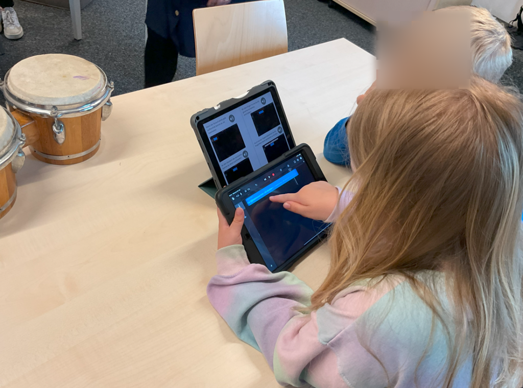 Zwei Kinder sitzen an einem Tisch und arbeiten an Tablets. Ein Kind zeigt mit dem Finger auf den Bildschirm, während das andere Kind zuschaut. Die Szene zeigt, wie die Kinder die Tablets zur Bearbeitung von Audio verwenden und diese zuschneiden.
