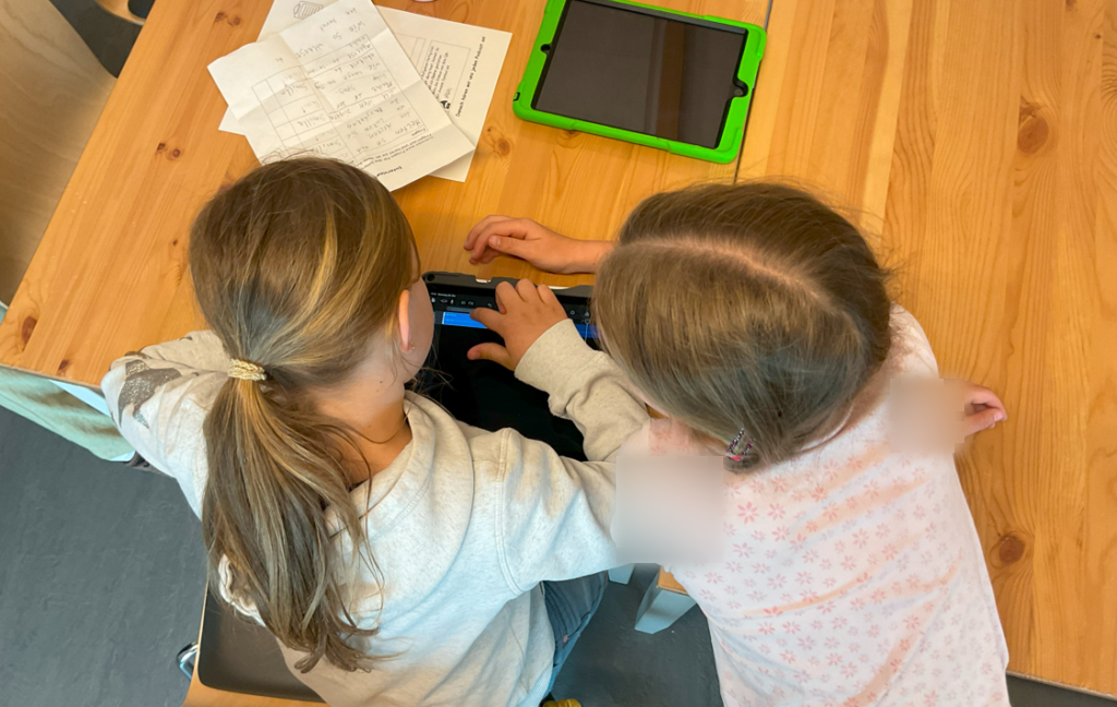 Zwei Mädchen sitzen an einem Holztisch und arbeiten gemeinsam an einem Tablet. Eines der Mädchen zeigt auf den Bildschirm, während das andere zuschaut. Auf dem Tisch liegen auch einige Papiere. Die Szene vermittelt konzentriertes und gemeinsames Arbeiten.
