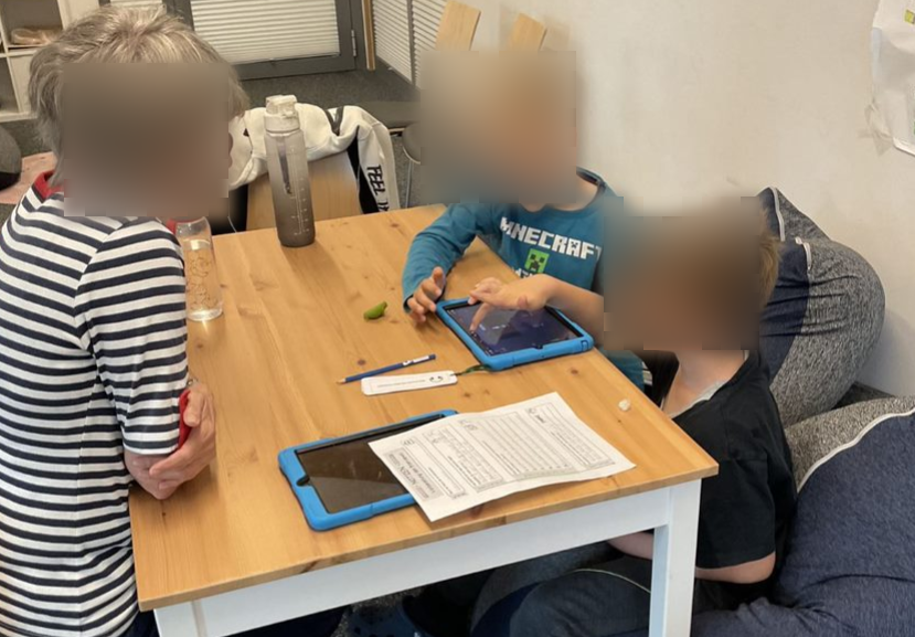 Zwei Jungen sitzen an einem Holztisch und arbeiten an einem Tablet. Einer der Jungen zeigt mit dem Finger auf den Bildschirm, während der andere aufmerksam zuschaut. Eine ältere Frau mit grauem Haar, die eine gestreifte Bluse trägt, steht neben dem Tisch und beobachtet die Jungen. Auf dem Tisch liegen ein weiteres Tablet, ein Bleistift und ein Arbeitsblatt.