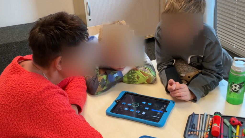 Zwei Jungen und eine Frau sitzen an einem Tisch und schauen konzentriert auf ein Tablet, das in der Mitte des Tisches liegt. Die Frau trägt einen roten Pullover und sitzt links, die Jungen tragen bunte Pullover und sitzen rechts.