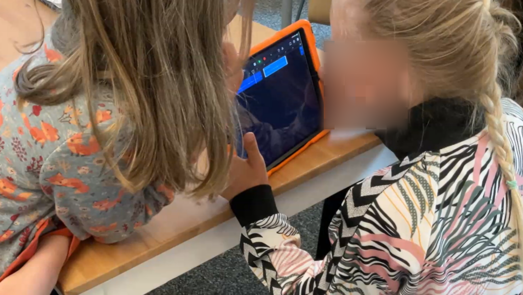 Zwei Mädchen sitzen nebeneinander an einem Tisch und arbeiten gemeinsam an einem Tablet mit einem orangefarbenen Rahmen. Sie sind konzentriert und schauen auf den Bildschirm, während sie miteinander interagieren. Die eine trägt eine Jacke mit schwarz-weißen und rosa Mustern, die andere trägt einen grauen Pullover mit bunten Mustern.