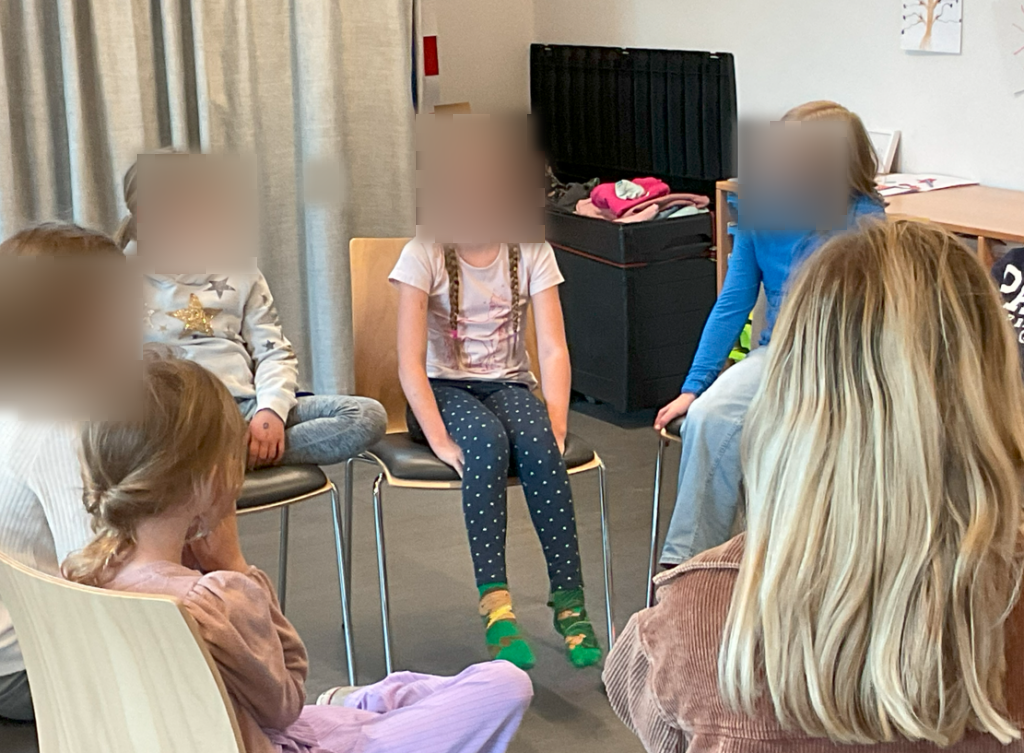 Eine Gruppe von Kindern sitzt im Kreis auf Stühlen und unterhält sich. Ein Mädchen in der Mitte lacht, während die anderen aufmerksam zuhören. Die Szene zeigt eine Reflexionsrunde, in der die Kinder ihre Erfahrungen teilen.