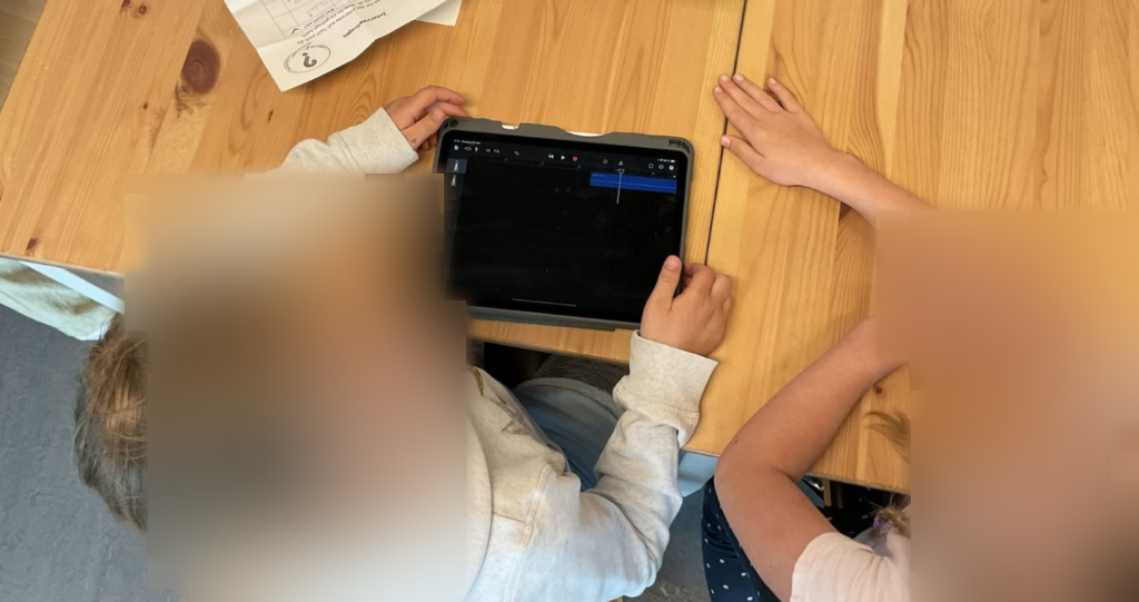 Zwei Mädchen sitzen nebeneinander an einem Holztisch und bearbeiten etwas auf einem Tablet. Sie sind konzentriert und scheinen eine Aufgabe gemeinsam zu lösen. Die beiden reden miteinander.