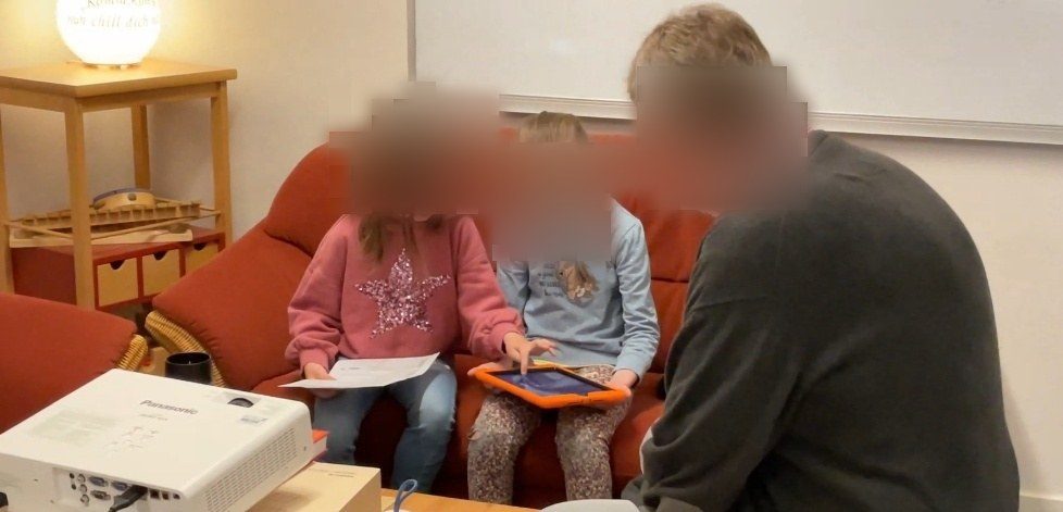 Auf dem Bild sind zwei Grundschulkinder in einem Klassenraum zu sehen, die auf einem roten Sofa sitzen. Sie führen ein Interview mit einem älteren Mann, vermutlich dem Großvater eines der Kinder. Eines der Kinder hält ein Blatt Papier in der Hand, während das andere Kind ein iPad benutzt, um die Antworten des Großvaters aufzunehmen. Der Großvater sitzt ihnen gegenüber und trägt ein dunkles Oberteil mit roten Streifen am Kragen. Der Raum ist gemütlich eingerichtet, mit einem Beistelltisch, auf dem ein Projektor steht, und einem Regal im Hintergrund. An der Wand hängt ein Whiteboard, und eine Lampe auf dem Tisch verbreitet ein warmes Licht. Die Atmosphäre wirkt ruhig und konzentriert, während die Kinder das Interview führen.