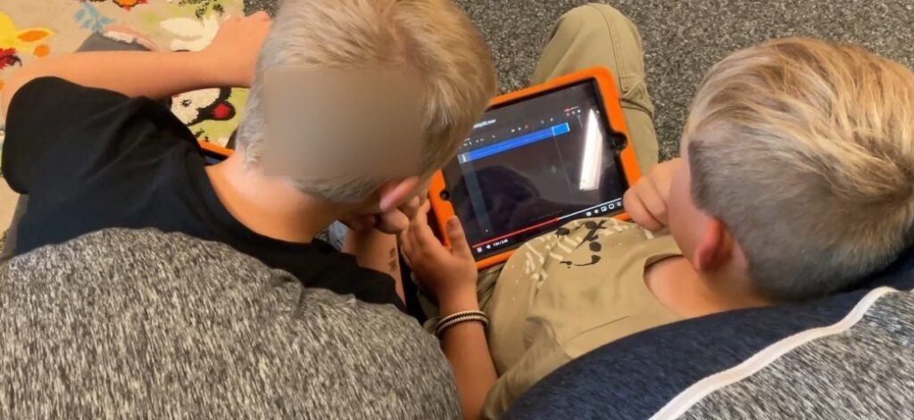 Auf dem Bild sind zwei Grundschulkinder zu sehen, die eng nebeneinander auf dem Boden sitzen und gemeinsam auf ein iPad schauen. Sie benutzen die App GarageBand, um eine Audiodatei zu bearbeiten. Das iPad hat eine orangefarbene Schutzhülle, und auf dem Bildschirm ist die Benutzeroberfläche der App zu erkennen, die das Schneiden von Audiodateien ermöglicht. Beide Kinder haben blonde Haare; eines trägt ein schwarzes Oberteil, das andere ein beigefarbenes T-Shirt mit einem Aufdruck. Der Boden ist mit einem grauen Teppich bedeckt, und im Hintergrund ist ein bunter Teppich mit Blumen- und Tiermustern zu sehen. Die Atmosphäre wirkt konzentriert, während die Kinder an ihrem Projekt arbeiten.