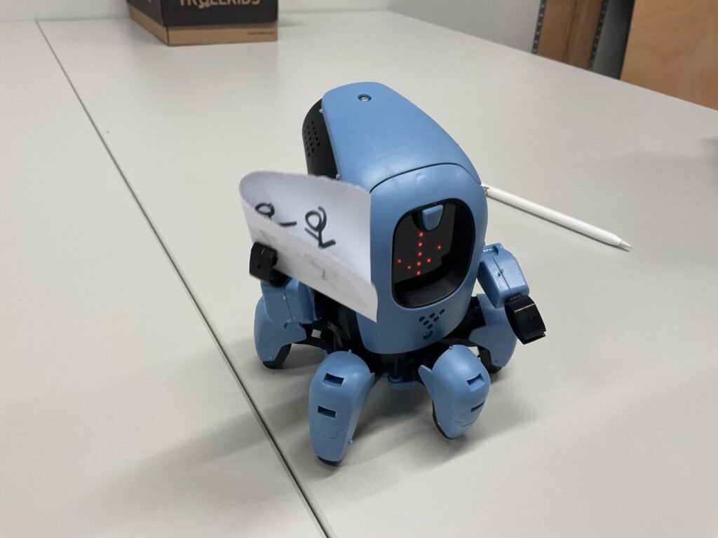 ein blauer Robotoer mit Spinnenbeinen und einem Display mit einem Pfeil drauf ist zu sehen