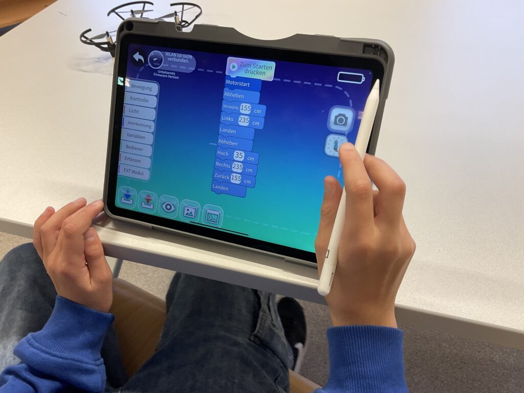 Auf dem Bild ist ein iPad zu sehen mit einer App in Blockprogrammiersprache. Der schüler hat einen Apple Pencil und tippt auf das iPad.