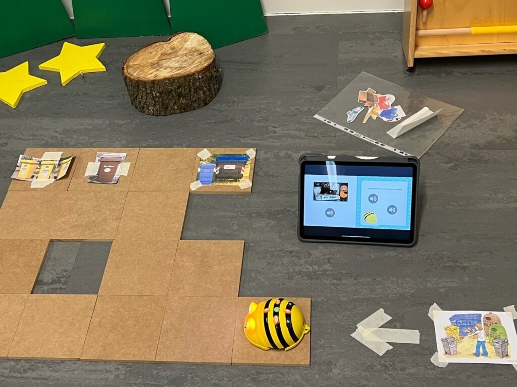 Station 1 Mülltrennung: Holzplatten auf dem Boden, BeeBot am Startpunkt, 3 Mülltonnen, ausgedruckter Müll und ein iPad.