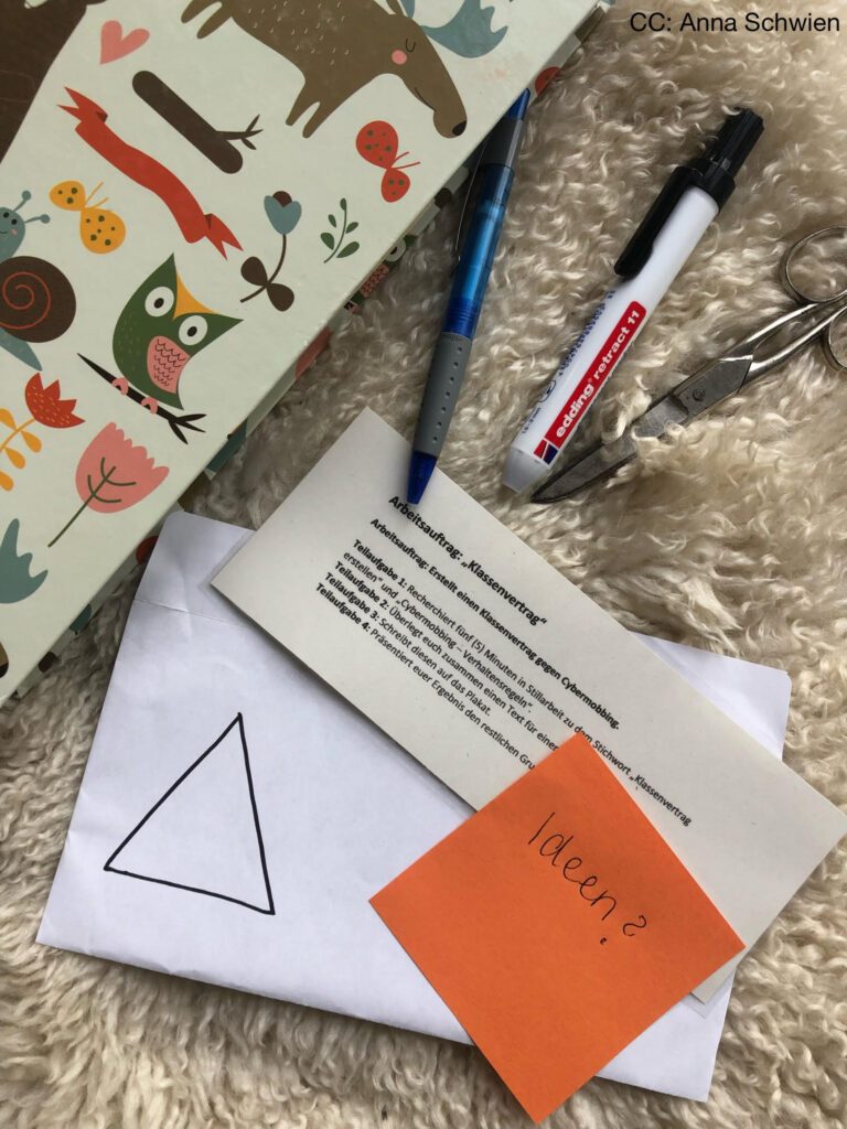 Der laminierte Arbeitsauftrag liegt neben einem geöffneten Briefumschlag mit dem Symbol Dreieck, Stiften, einer Schere und einem orangenem Post It mit der Aufschrift "Ideen?"